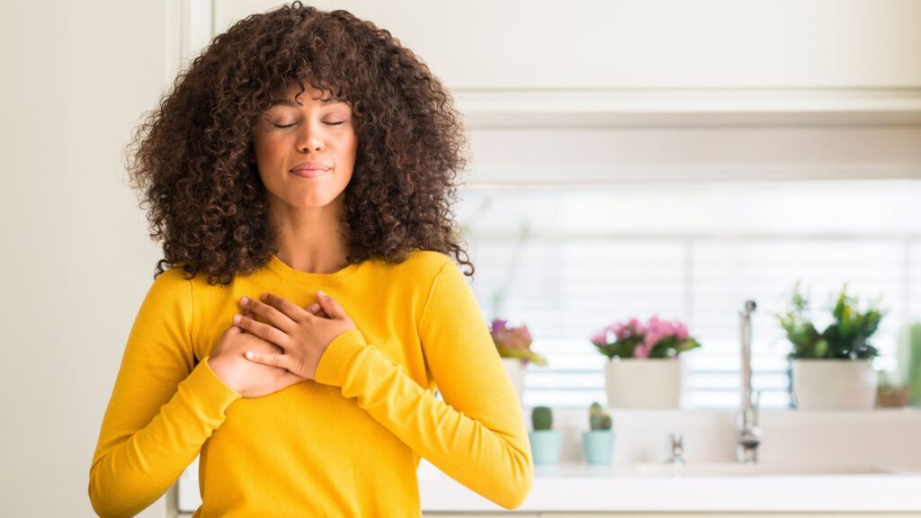 African american woman wearing yellow sweater at kitchen smiling with hands on chest with closed eyes and grateful gesture on face.