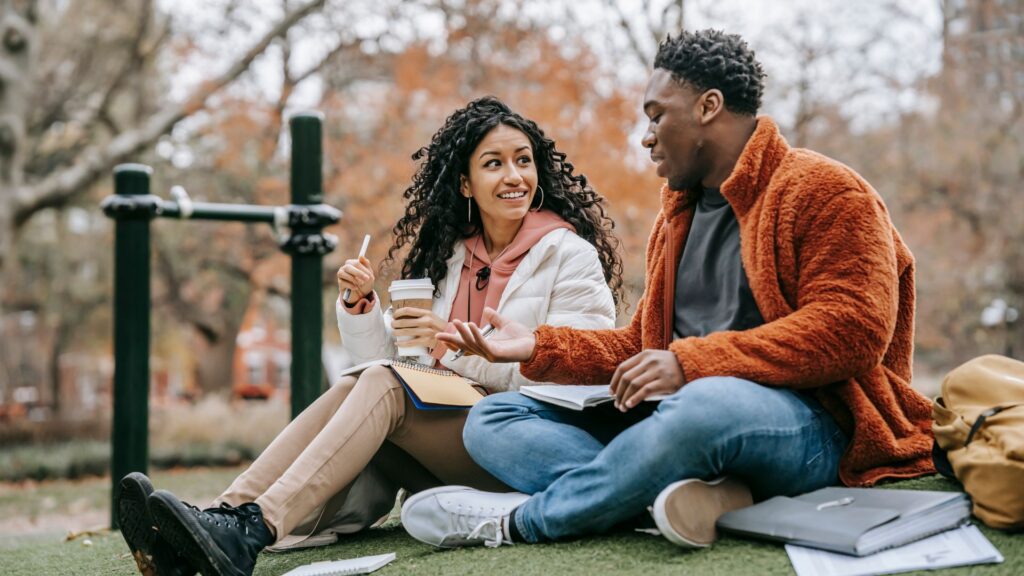 Multiethnic couple with notebooks communicating on grassy lawn while studying together